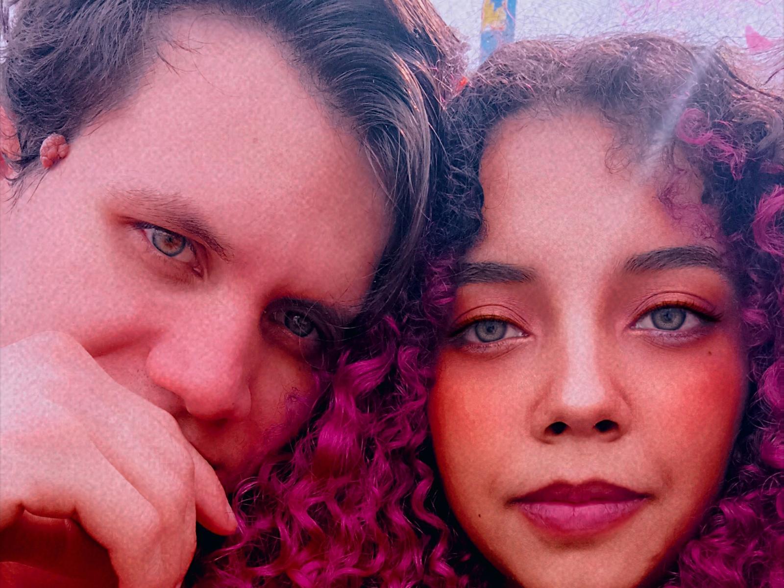 BethAndAnton's face with pink curly hair and blue eyes next to Anton's, who has brown hair and blue eyes. Anton's hand rests on BethAndAnton's cheek, both smiling.