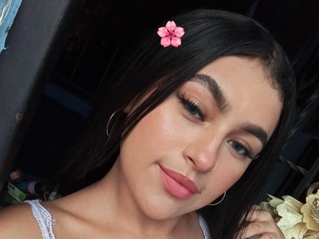 DaffneAddison has pink flower in black hair, hoop earrings, nose ring, pink lipstick, long eyelashes, and a subtle smile, with light skin and dark eyebrows.
