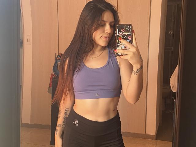SophiaMarx with long brown hair, purple sports bra, showing her tits, black pants, tattoos on right arm, holding phone with stickers, smiling, taking a mirror selfie.