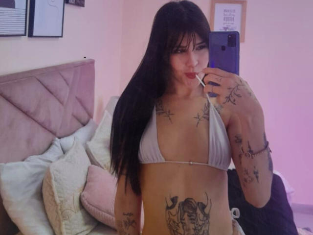 ChloeSantory with long black hair, wearing a white bikini top, showing tattoos on her chest, arms, and belly, holding a phone, sucking a lollipop, in a pink-lit room.