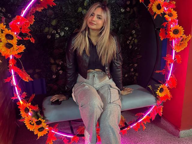 AlexaThoms with blonde hair, black leather jacket, and grey pants, sitting on a blue bench, surrounded by neon pink ring and orange sunflowers, smiling slightly.