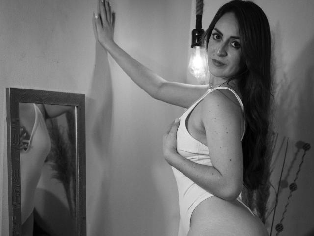 ButtyCherry with long hair, wearing a white tank top, covering her tits, leaning against a wall, smiling, standing next to a mirror with a light bulb above.