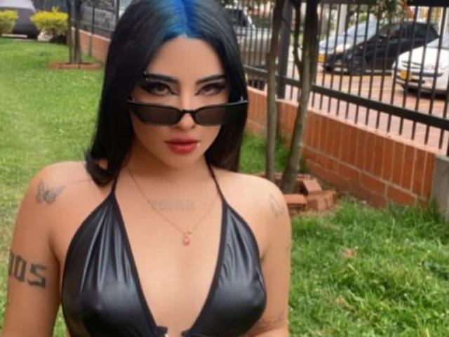 NatallieBlue with black hair, blue highlights, wearing a deep black leather halter top, black sunglasses, tattoos on shoulders, and a necklace, standing outside on a grassy lawn.