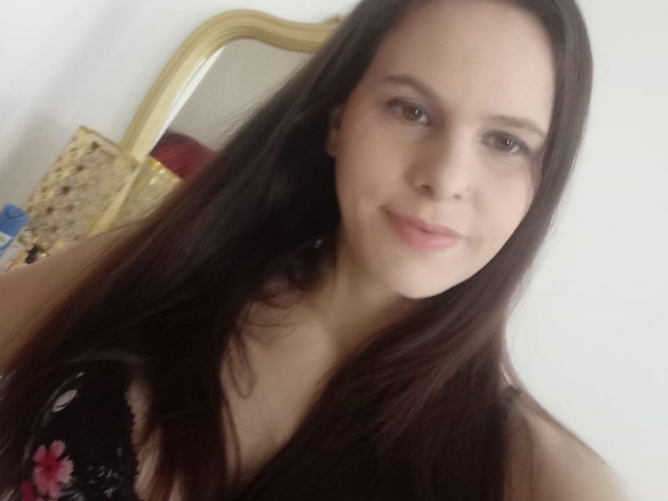 MimiXo has long brown hair, light skin, and a slight smile with pink lips. She's wearing a black floral dress, showing her small tits and a cute face.