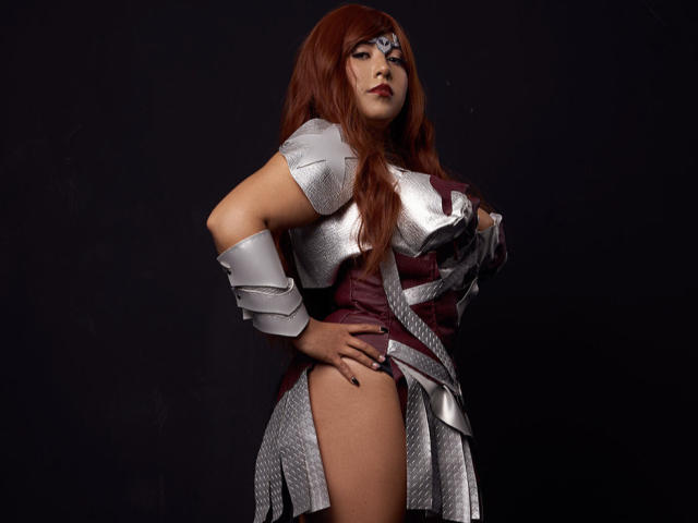 EvangelineAx with long red hair, wearing a silver armored top and maroon skirt, showing her bare ass, hand on hip, dark background, looking confident.