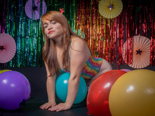 MoonRay69 met rood haar, een gemiddelde borstmaat en een kleurrijke tie-dye badpak, knielt op handen en voeten, omringd door kleurrijke ballonnen, en kijkt met een lichte glimlach naar de camera..