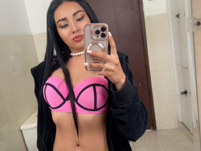 LillyTaylor with long black hair, pink bra with black lines, showing her tits, wearing a black jacket, holding a phone, pearl necklace, in a bathroom.