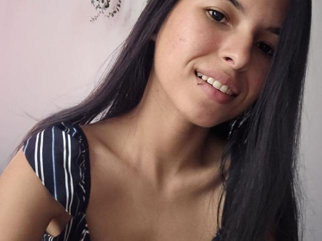 JuliaLanne with long black hair, smiling, showing teeth, wearing a striped blue and white top, has a small nose piercing, and has her tits slightly visible in the frame.