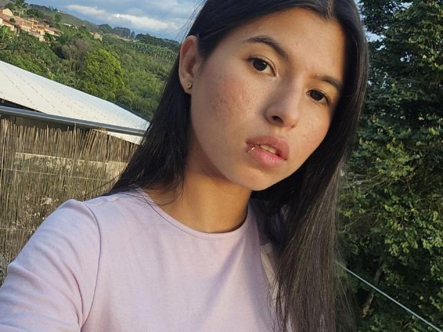 JuliaLanne has medium brown skin, long black hair, and is wearing a light pink shirt. Her small tits are visible. Her face has freckles and she's looking at the camera.