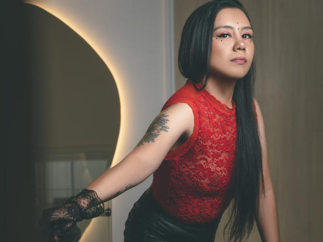 CharlotteJoliie with long black hair, red lace top, black leather skirt, tattoo on right arm, black lace glove, and a facial dot in the center. She's leaning forward.