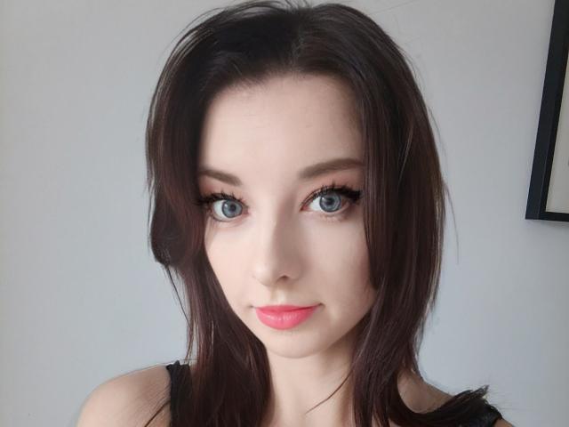 DominaLuna has blue eyes, long brown hair, and pink lipstick. She's wearing black eyeliner and has a cute, slightly smoky look. Her tits are not visible.