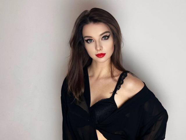 DominaLuna with red lipstick, dark brown hair, black lace bra, and black sheer robe, exposing her tits, standing against a white wall, looking directly at the camera.