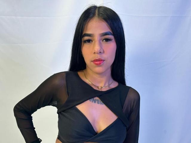 EmilyLuzz has long black hair, wearing a black sheer top with a cutout that shows her tits, red lipstick, and a nose ring, standing against a white background.