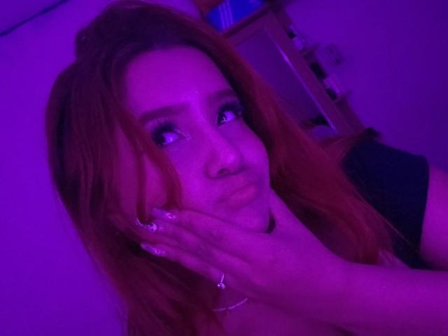 AlanaOdinsson with pinkish-purple lit face, hand touching cheek, long red hair, looking to the side, wearing a black top, ring on finger, nail art, slight pout.