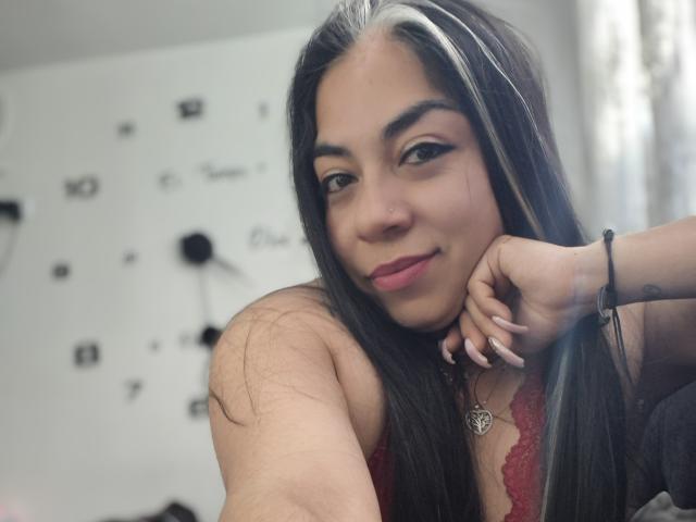 EvaLunaLatina with long black hair, white highlights, and pink lipstick, wearing a red lace top, smiling at the camera, resting her chin on her hand.