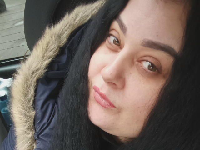 EleanorViski with dark hair, brown eyes, and full pink lips, wearing a black coat with a fur hood, looking slightly to the side with a subtle smile.
