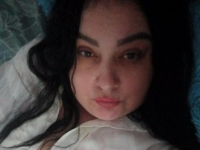 EleanorViski's face, dark wavy hair, brown eyes, and pink lips, wearing a white shirt, lying down, soft lighting, subtle shadow on her right cheek.