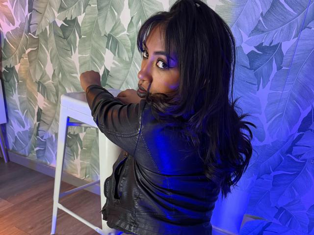 BlackXRose with long black hair, looking over her shoulder, wearing a black leather jacket, touching a white table, standing on wooden floor, blue light in background.