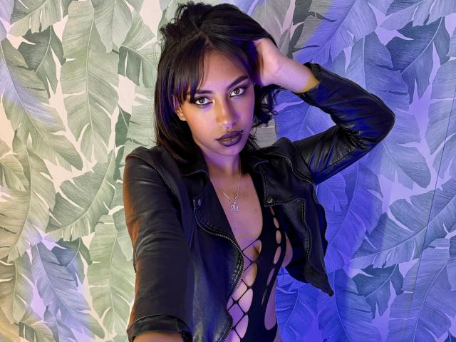 BlackXRose with dark skin, black leather jacket, and lattice-style black top showing her tits. She has purple lipstick, black hair, and a hand in her hair. Blue lighting.