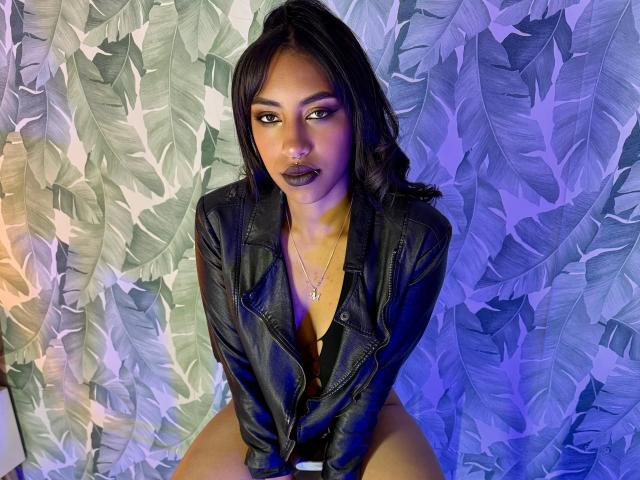 BlackXRose with dark lipstick, black leather jacket, and necklace, sitting with legs apart, looking up with intense eyes, wearing black lingerie, blue and purple lighting.