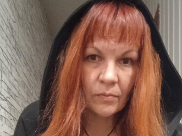 AugieCherry with red-orange hair, black hooded jacket, and serious expression, showing her nose and slightly puffed lips. She has a fair skin tone and bangs.