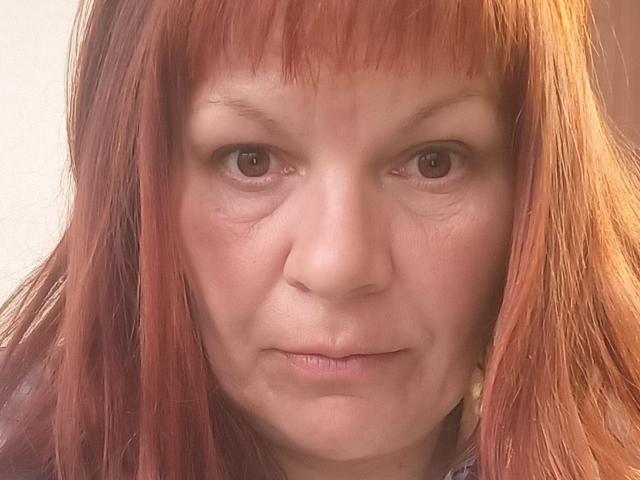 AugieCherry has red hair, brown eyes, and a serious expression. Her face shows slight wrinkles and she's wearing a pearl earring on the right side.
