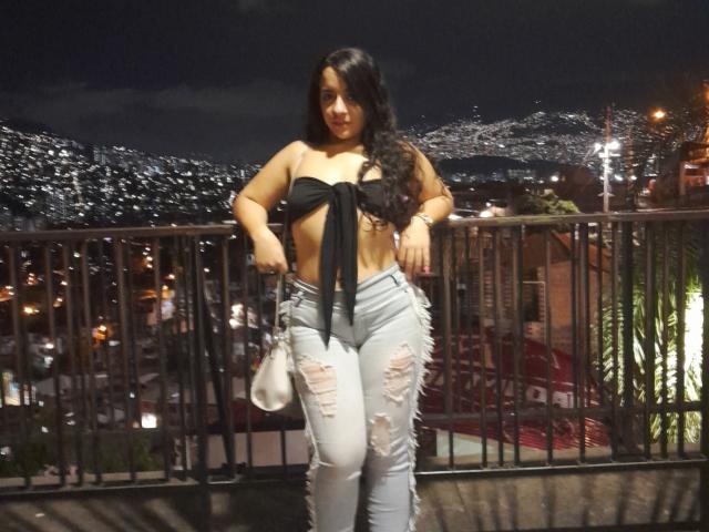 LindaKurlin with long black hair, wearing a black strapless top tied in the front, and ripped light blue jeans, standing on a balcony at night, holding a white bag.