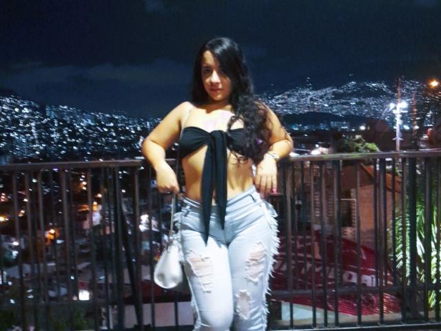 LindaKurlin with long black hair, wearing a black tied-up top exposing her boobs and ripped light blue jeans, standing on a balcony at night, holding a white bag.
