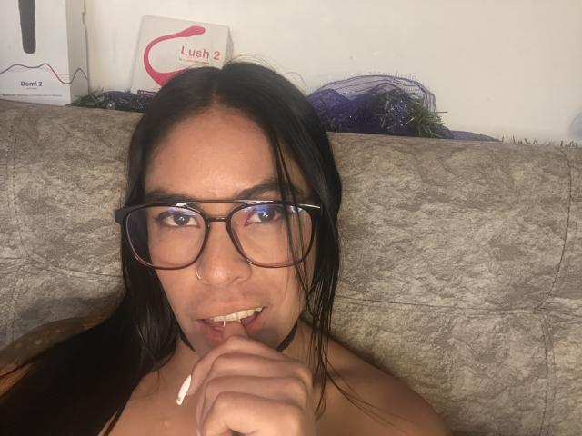 StellaBeckker with black hair, glasses, nose ring, and long nails, biting her finger, looking into the camera, sitting on a gray couch.
