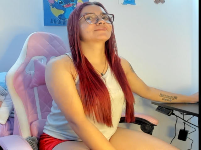 EimmyHoskX with red hair, glasses, white tank top, red shorts, sitting on pink and white gaming chair, tattoo on right forearm, looking up, slight smirk, arm on desk.