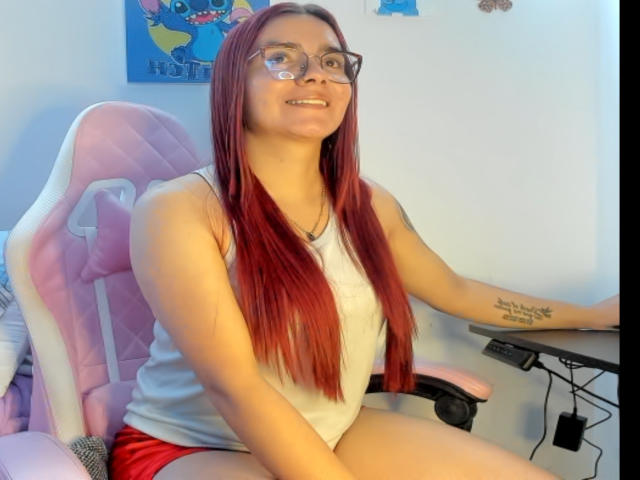 EimmyHoskX with red hair, glasses, white tank top, red shorts, pink gaming chair, tattoo on right arm, smiling, sitting, right hand on desk, left arm resting.