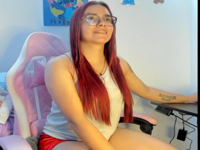 EimmyHoskX with long red hair, glasses, white tank top, red shorts, sitting on pink gaming chair, tattoo on right arm, smiling, hands on black desk.