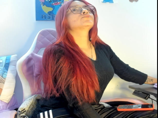 EimmyHoskX with long red hair, glasses, black top, black pants, sitting in a chair, right hand on a desk, left arm tattooed, looking up, slightly pouty expression.