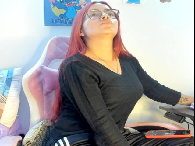 EimmyHoskX, red-haired woman with glasses, black V-neck, sitting on pink and white chair, looking up, tattoo on right wrist, black and white striped pants, holding a flat surface.