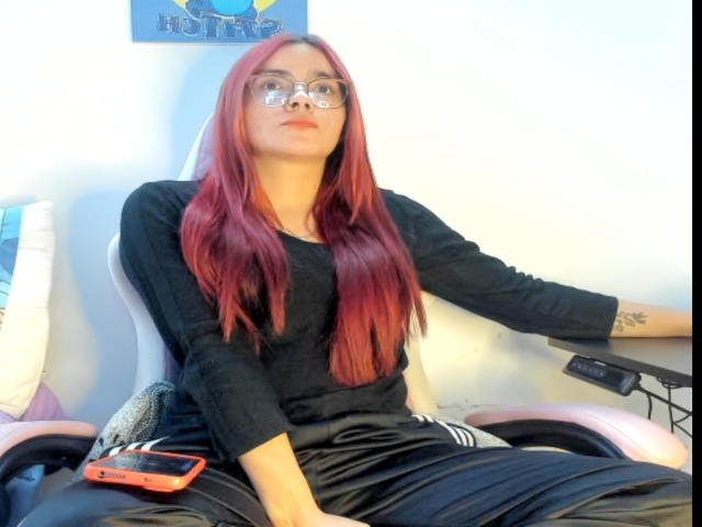 EimmyHoskX with red hair, glasses, black long-sleeve shirt, black pants, sitting on a pink chair, phone on left, arm tattoo, white wall, slightly pouty face.