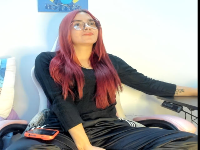 EimmyHoskX with red hair, glasses, black shirt, and black pants, sitting in a chair, pouting, with a red phone on the armrest.