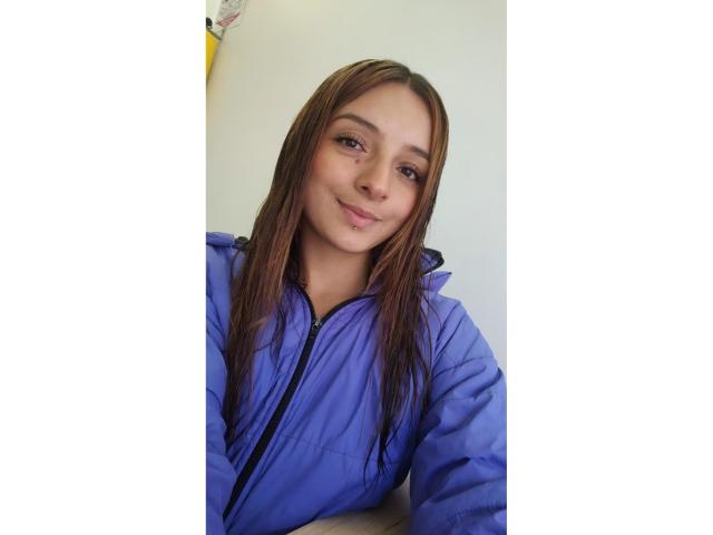 SaarahJones with long brown hair, wearing a blue jacket, smiling slightly, her small tits barely visible under the jacket, white background, casual selfie.