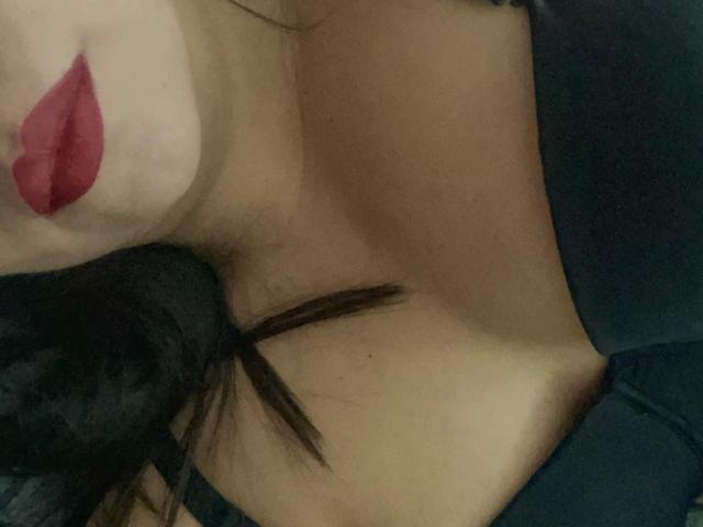 LilyRousee's face with red lipstick, black hair, and a visible cleavage in a dark top, showing her armpit and part of her chest.