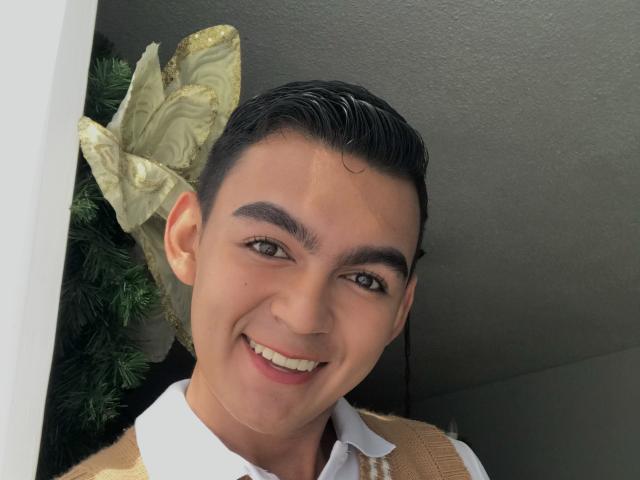 DeusBeauty, smiling with dark slicked-back hair, wearing a white shirt and beige vest, has brown eyes and a cute face. A large yellow bow is behind him.