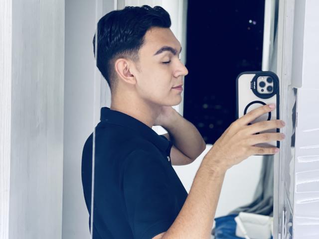 DeusBeauty with short black hair, closed eyes, and a black shirt, taking a selfie in a mirror using a smartphone with a black case.