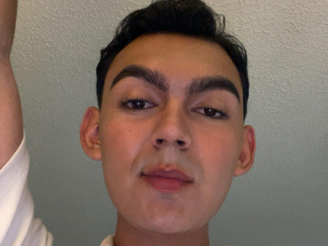 DeusBeauty has dark, thick eyebrows, brown eyes, and a slight smile with pink lips. Short black hair, light skin, and a white shirt. Textured ceiling in background.