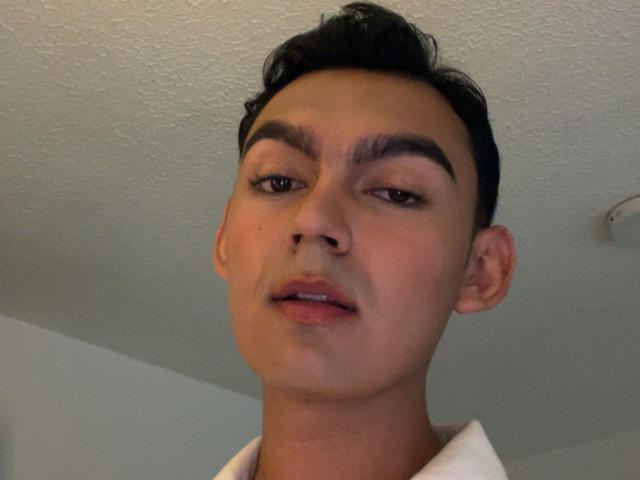 DeusBeauty has a slim face with dark, thick eyebrows, short black hair, and a slightly open mouth with pink lips. White ceiling in the background.