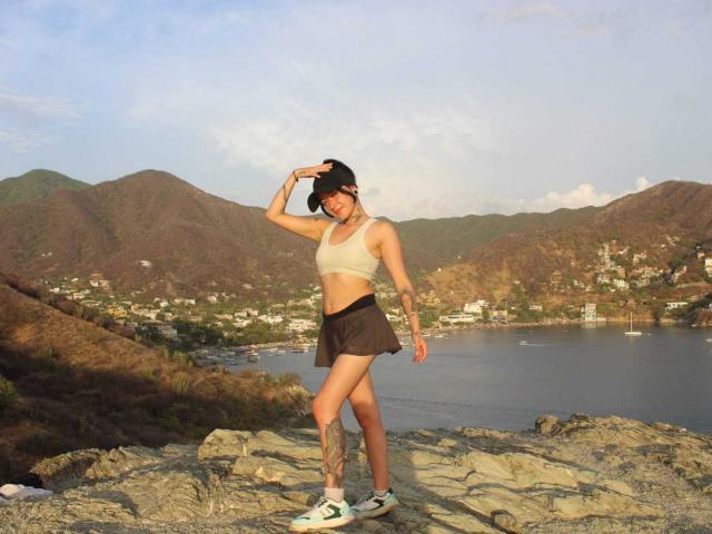 AriannaWhiteX with a slim build, small tits, tattoo on left thigh, wearing a beige sports bra, black skirt, white sneakers, standing on rocky terrain, hand in hair.
