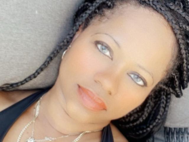 Cincia69 a la peau brun clair, les yeux bleus et de petites lèvres orange. Elle a les cheveux noirs tressés, porte un haut noir et un collier en argent, et est allongée avec une expression neutre..