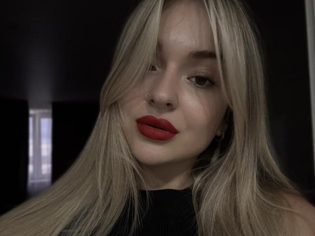EvaAdderiy with long blonde hair, red lipstick, nose ring, and black sleeveless top, looking slightly to the side, has a soft smile and light skin.