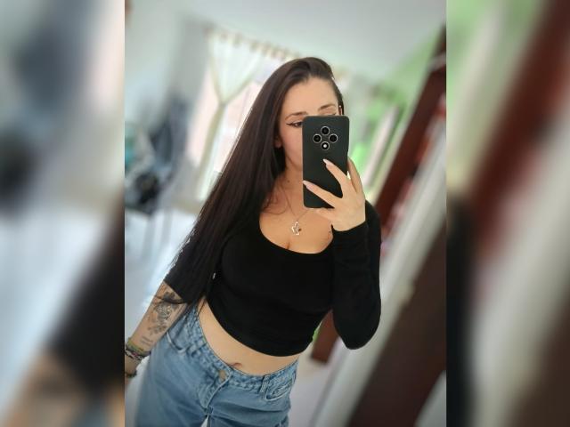 CanellaDiaz, met lang zwart haar, een zwarte crop top en blauwe jeans, houdt een zwarte telefoon vast, toont haar getatoeëerde arm en draagt een ketting met een hartvormige hanger..