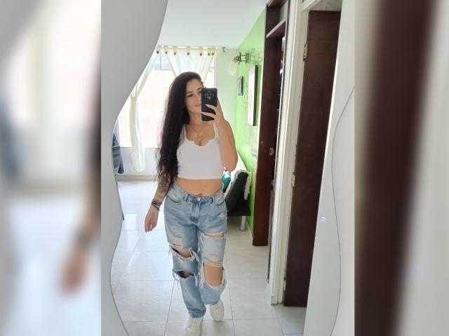 CanellaDiaz, con lunghi capelli scuri, un top bianco a vita corta e jeans blu strappati, sta scattando un selfie allo specchio, mostrando il seno e i glutei, in piedi in una stanza con piastrelle bianche..