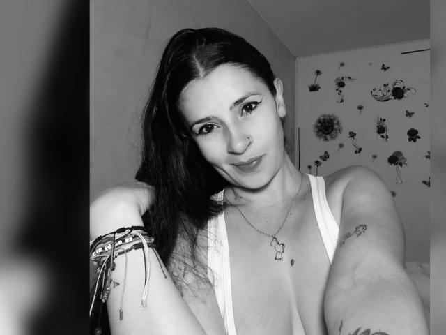 Black-and-white selfie of CanellaDiaz with long dark hair, nose ring, and bracelet, wearing a low-cut top, showing small tits, tattoos on arms, and a necklace. Smiling.