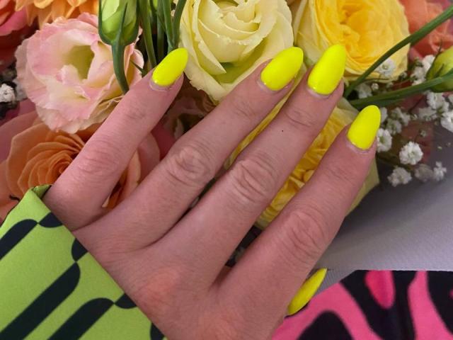 AlicePrings' hand with bright yellow neon nails, fingers slightly spread, showing off long, sharp, glossy nails against a bouquet of roses. Her skin is fair, and she's wearing green.