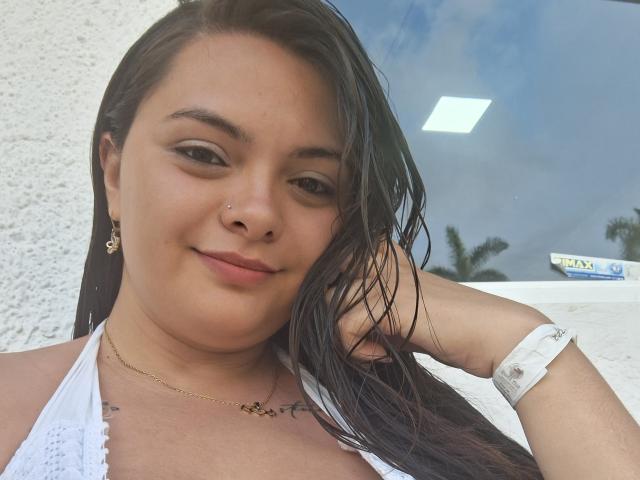 Katherin Latin heeft nat zwart haar, een neusring en een kleine tatoeage op haar borst. Ze draagt een witte top met halter, glimlacht licht en heeft een witte polsband om..